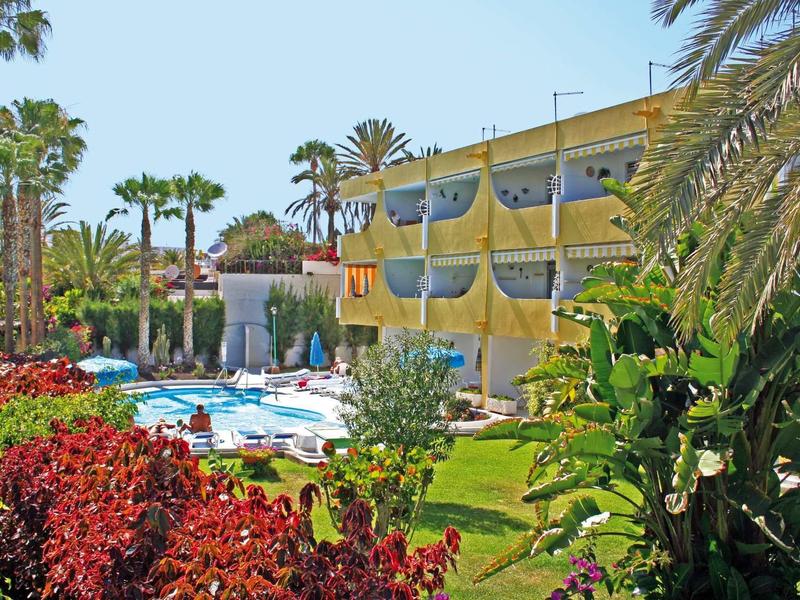 Hotel mit gelbem Gebäude, blauem Pool, grünen Palmen und bunten Blumen im Garten.