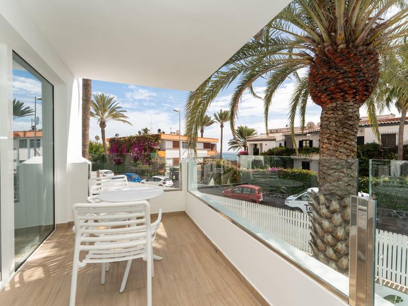 Balkon z krzesłami i stołem, palmy i domy w tle pod czystym niebem.