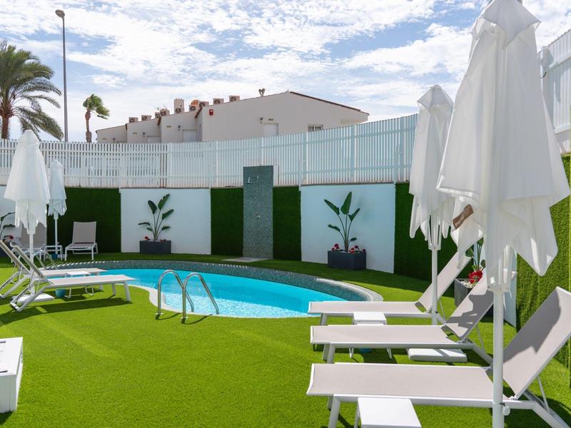 Strefa przy basenie z leżakami, parasolami i sztuczną trawą pod niebieskim niebem.