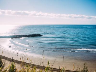 Breiter Sandstrand mit sanften Wellen und wenigen Menschen, dahinter ruhiges blaues Meer.