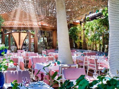Offenes Restaurant mit rosafarbenen Tischen, weißen Stühlen und grünen Pflanzen unter Baumkronen.