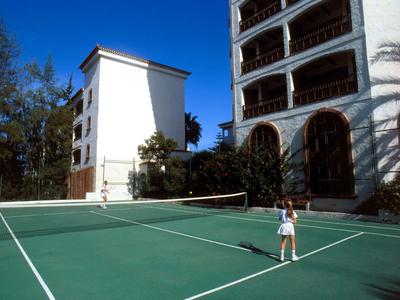 Outdoor-Tennisplatz mit grünem Boden, zwei Gebäuden und einer spielenden Person in weißer Kleidung.