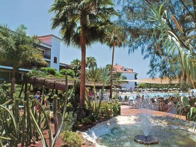 Hotelanlage mit Pool, Palmen, grünen Pflanzen und blauen Himmel im Hintergrund.