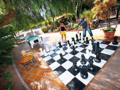 Zwei Personen spielen Schach auf großem Brett im Garten mit Pflanzen und Terrakottaboden.