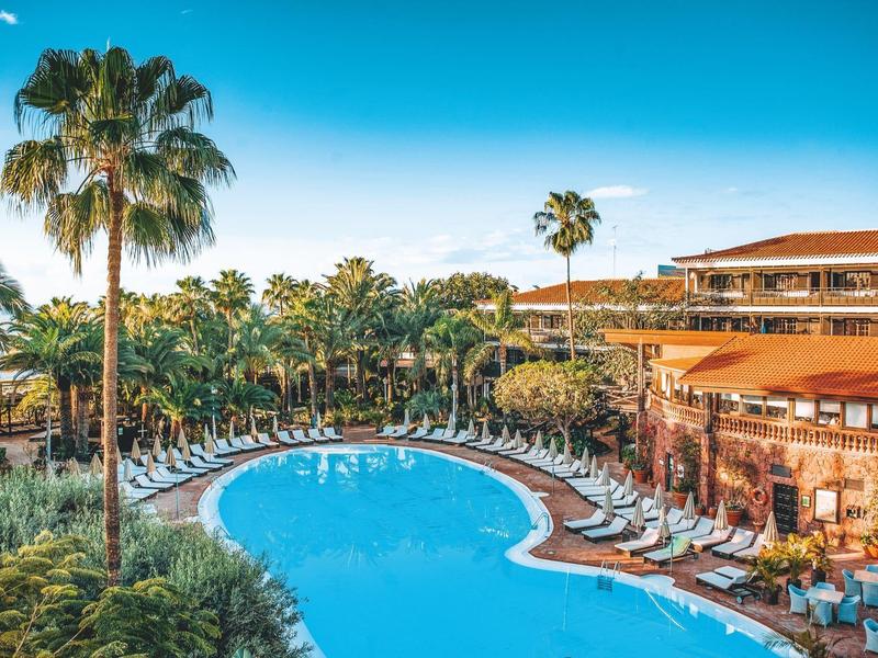 Duży basen hotelowy otoczony palmami i leżakami pod jasnym niebieskim niebem.