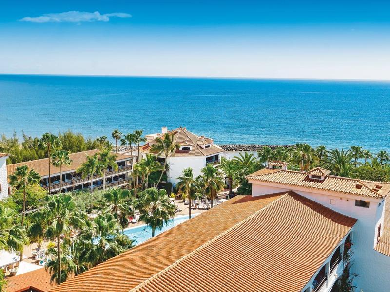 Hotel z czerwonymi dachami, palmami i basenem nad morzem pod niebieskim niebem.