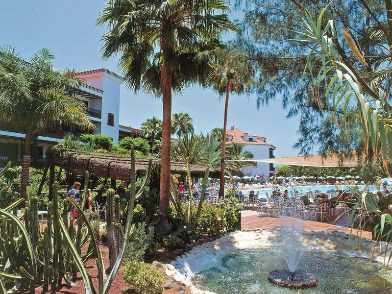 Ogród hotelowy z palmami, basenem, zadaszonymi miejscami do siedzenia i fontanną pod bezchmurnym niebem.