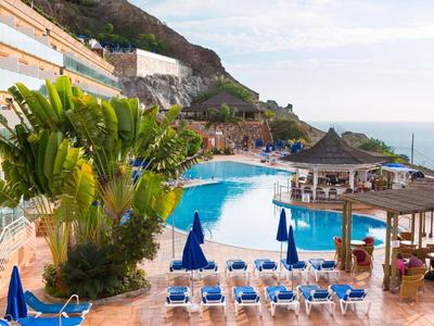 Zwembad van resort met blauwe ligstoelen, palmbomen en uitzicht op oceaan bij heuvels.