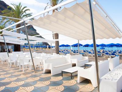 Buiten zitgedeelte met witte banken en tafels onder een luifel bij een strand met blauwe parasols.