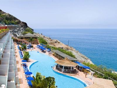 Terras met zitplaatsen bij een hotel met uitzicht op een gebogen zwembad en blauwe oceaan onder een heldere hemel.