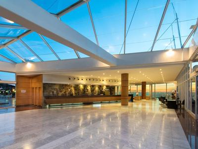 Ruime hotelhal met marmeren vloer, houten receptiebalie en glazen dak onder blauwe lucht.