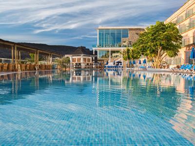 Duży basen na zewnątrz z niebieską powierzchnią z płytek otoczony leżakami i nowoczesną architekturą hotelu.