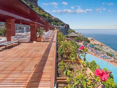 Terras aan zee met houten vloer, ligstoelen, bloemen, zwembad en uitzicht op oceaan bij blauwe hemel.