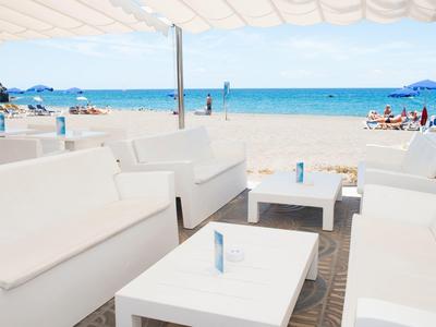 Wit beklede buitenzitplaatsen met tafels onder schaduw, uitzicht op zonnig strand en kalme blauwe zee.