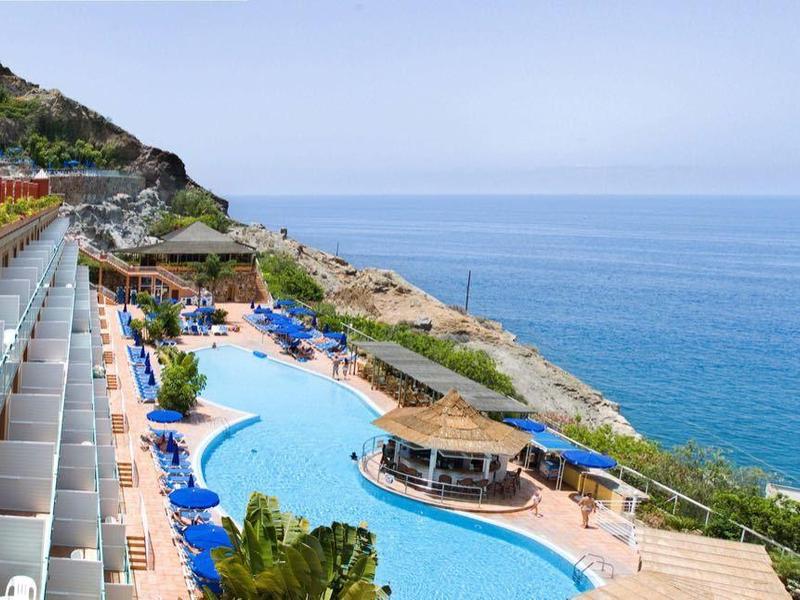 Terras met zitplaatsen bij een hotel met uitzicht op een gebogen zwembad en blauwe oceaan onder een heldere hemel.