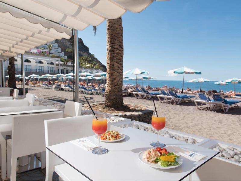 Strandrestaurant met witte tafels die drankjes en eten serveren bij ligbedden en parasols.