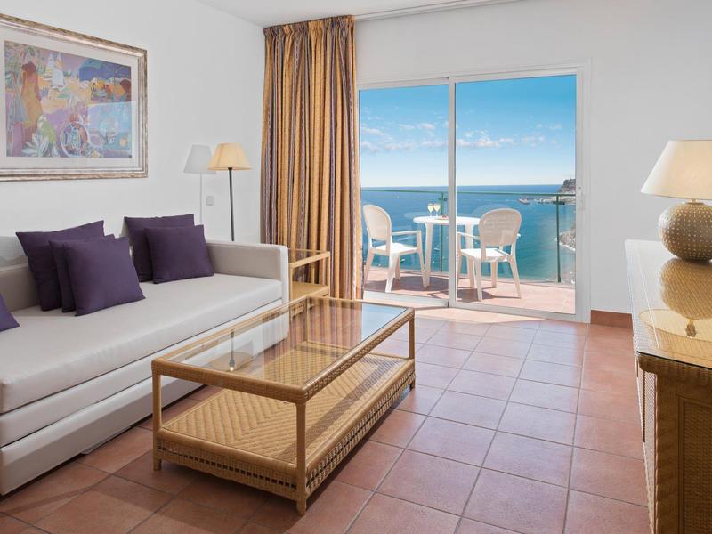 Lichte hotelkamer met bank, glazen salontafel en balkon met uitzicht op de oceaan.