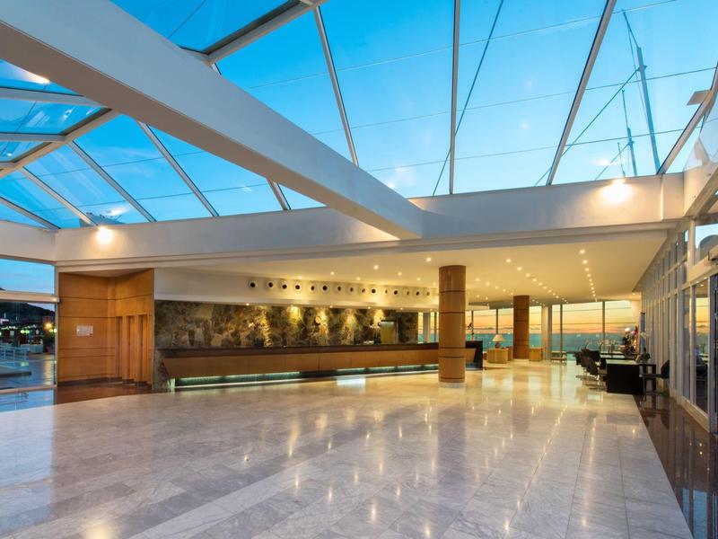 Ruime hotelhal met marmeren vloer, houten receptiebalie en glazen dak onder blauwe lucht.