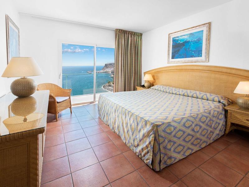 Lichte hotelkamer met betegelde vloer, tweepersoonsbed, houten meubels en raam met zeezicht.