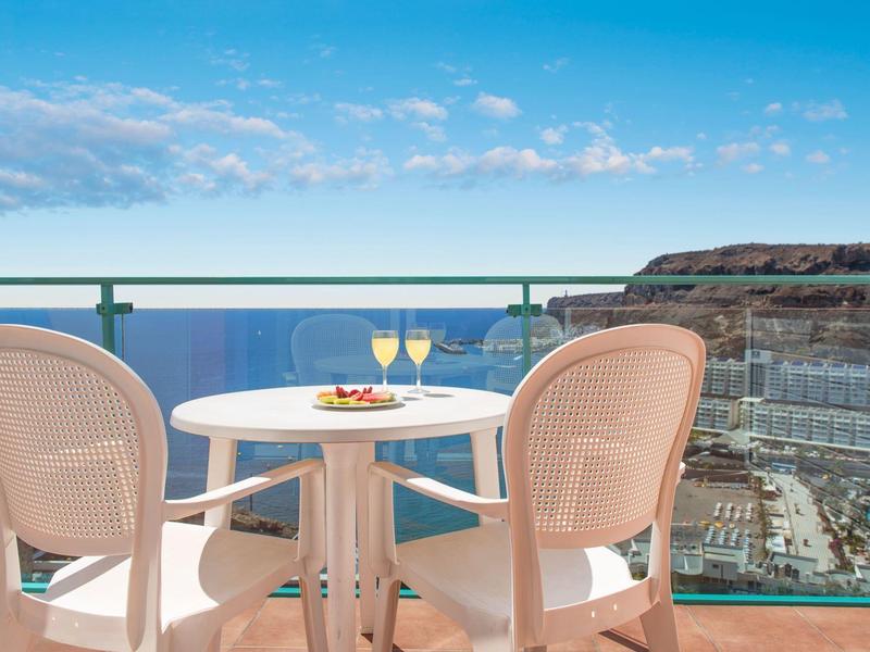 Balkon met tafel, twee stoelen, drankjes en uitzicht op de oceaan bij een zonnig hotel.