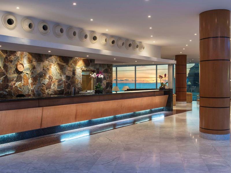 Ruime hotel lobby met moderne receptiebalie en uitzicht op de oceaan door grote ramen.