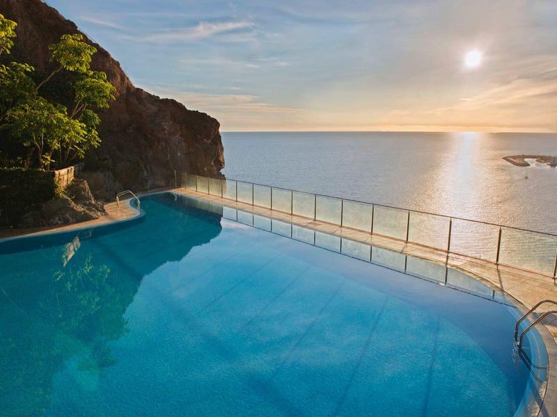 Infinity zwembad met uitzicht op de oceaan bij zonsondergang en een rotswand links.