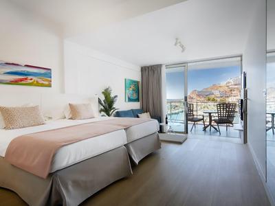 Bright hotel room with two beds, balcony, and view of coast and town.