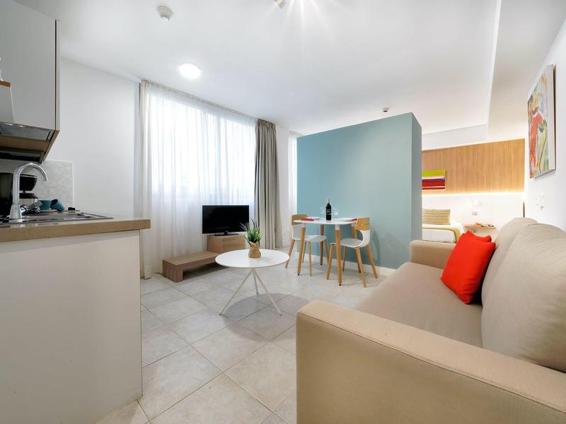 Salon lumineux avec canapé beige, petite table à manger et coin cuisine.