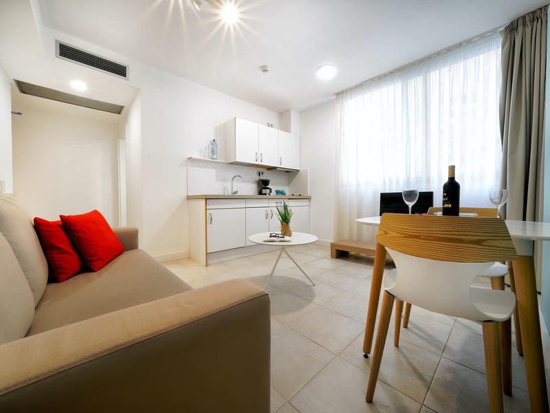 Chambre d'hôtel cosy avec canapé beige, coussins rouges, kitchenette et coin repas lumineux.