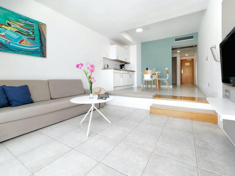 Salon moderne avec canapé gris, table basse blanche et petite cuisine en arrière-plan