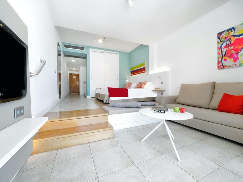 Chambre d'hôtel moderne avec lit, canapé, TV, coussins colorés et œuvre d'art au mur.