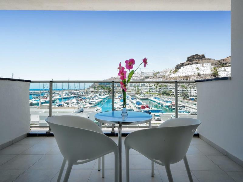 Balcon avec deux chaises et une table donnant sur une marina et une ville sur la colline sous un ciel clair.
