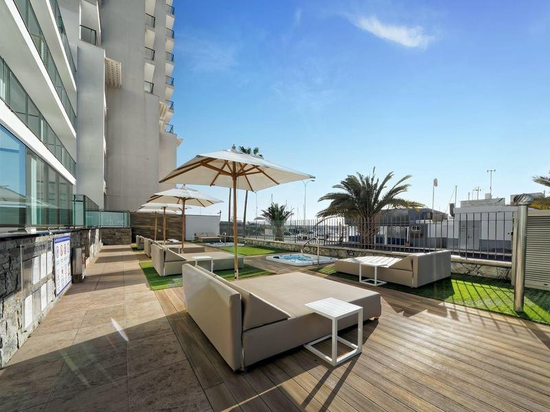 Espace lounge extérieur avec canapés, parasols, terrasse en bois et palmiers sous un ciel clair.