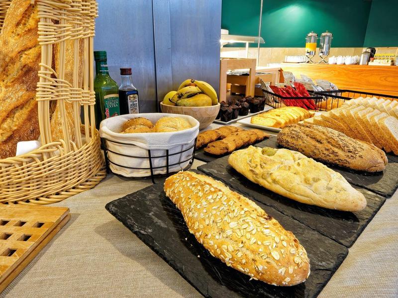 Variété de pains frais et petits pains présentés sur un comptoir avec des paniers et des fruits en arrière-plan.