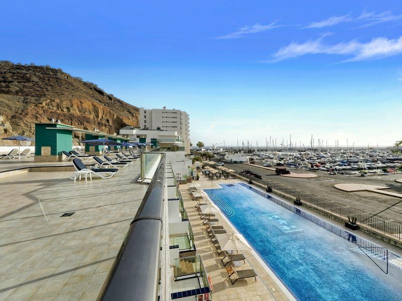 Piscine extérieure et marina d'un hôtel près de collines rocheuses sous un ciel bleu clair.