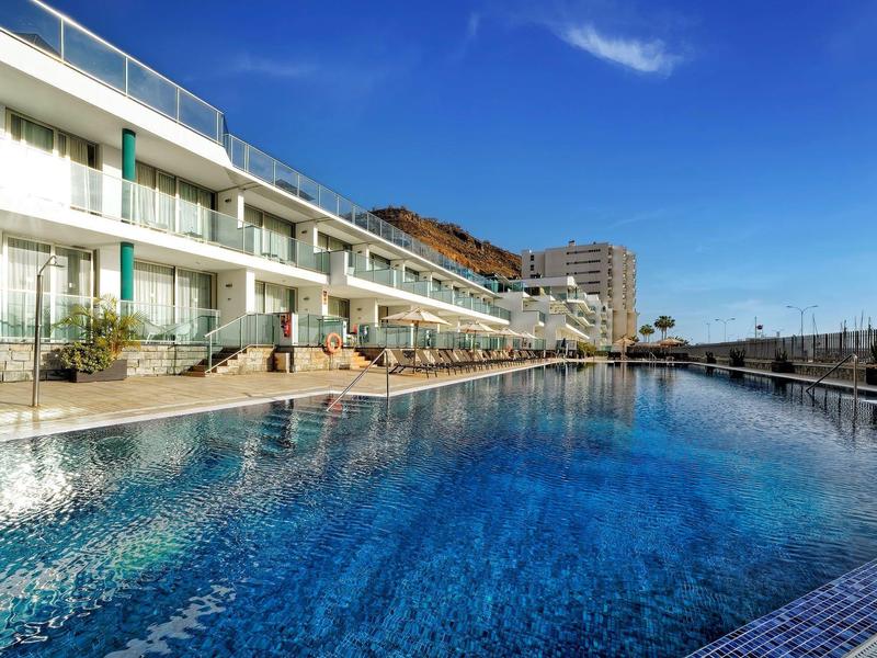 Hôtel moderne avec grande piscine extérieure et collines en arrière-plan sous un ciel bleu clair.