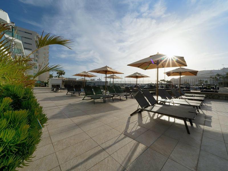 Terrasse sur le toit ensoleillée avec chaises longues, parasols et vue sur la ville sous un ciel partiellement nuageux.