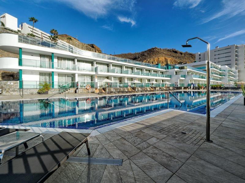 Hôtel moderne avec grande piscine extérieure et transats sous un ciel bleu.