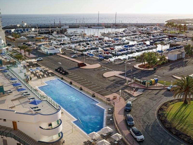 Station balnéaire ensoleillé avec une grande piscine, des chaises longues, des palmiers et une marina remplie de bateaux.