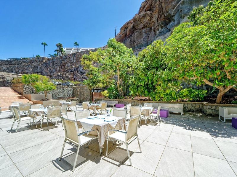 Terrasse extérieure avec tables et chaises sous ciel dégagé près d'une colline rocheuse et de verdure.