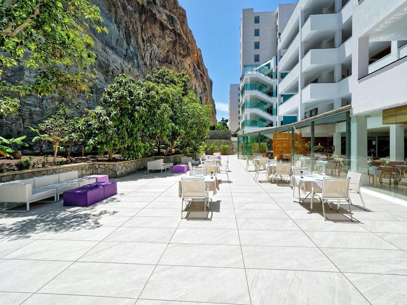 Grande terrasse d'hôtel avec carrelage blanc, chaises modernes et jardin au pied de la falaise.