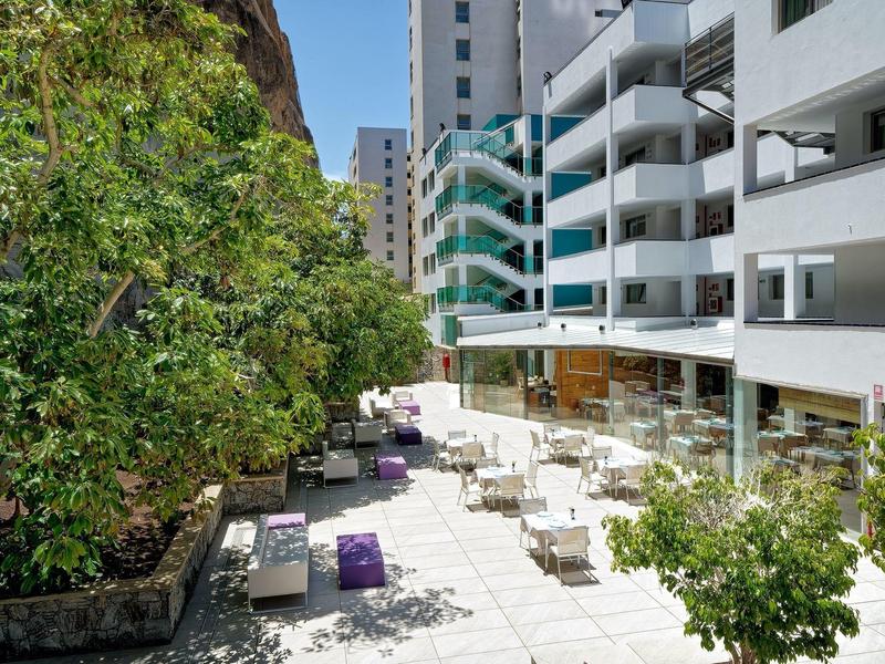 Terrasse moderne d'hôtel avec tables et chaises blanches entourées de verdure et de grands bâtiments.