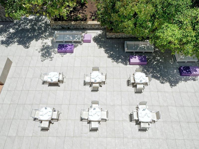 Espace extérieur avec six tables blanches et bancs, entouré d'arbres verts et de jardinières violettes.