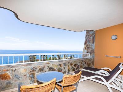Balkon mit Steinmauer, zwei Liegestühlen, Tisch, Stühlen und Meerblick bei klarem Himmel.