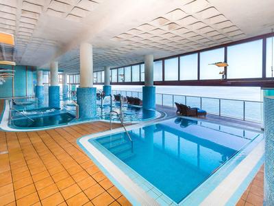 Innenraum mit zwei blauen Schwimmbecken, orangen Fliesen und großen Panoramafenstern.