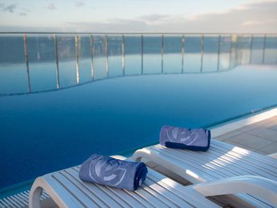 Moderner Infinity-Pool mit Glasgeländer und zwei weißen Liegestühlen mit blauen Handtüchern.