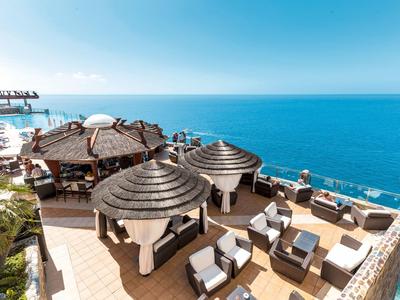 Terrasse mit runden Strohdächern, weißen Sesseln und Blick auf ruhiges blaues Meer.