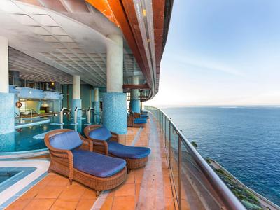 Balkon mit blauen Liegestühlen und Glasgeländer über dem ruhigen Meer bei klarem Himmel.