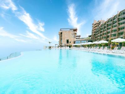 Infinity-Pool mit klarem Wasser, neben einem modernen Hotel und blauem Himmel mit Wolken.