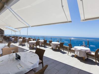 Terrassenrestaurant mit weißen Tischen und Sonnenschutz, Blick auf blaues Meer und Himmel.